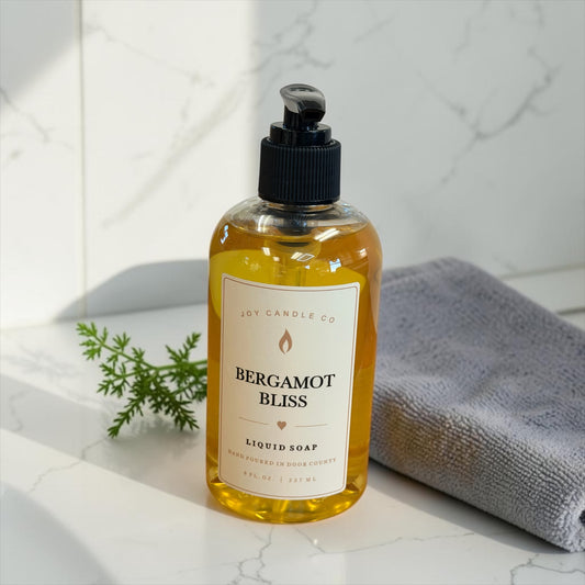 Bergamot Bliss liquid hand soap bottle on a white marble countertop with a folded gray towel and a sprig of greenery